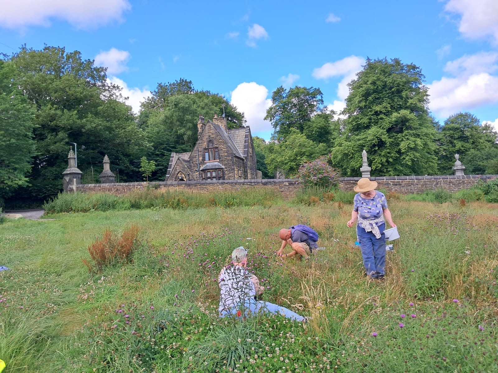 Three people working in a meadow with trees and a building behind
