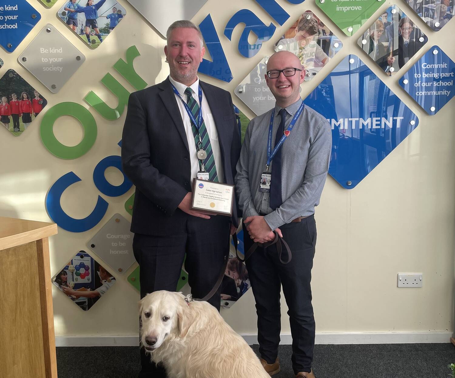 Two male teachers holding certificate with golden retriever dog in front of them
